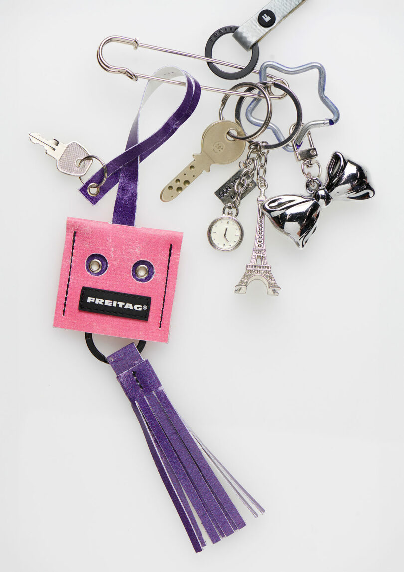 A set of keys and various keychains, including a pink FREITAG tag, Lucky Trucker Charms, a purple tassel, a silver Eiffel Tower, a bow, a watch charm, and a star-shaped keyring.