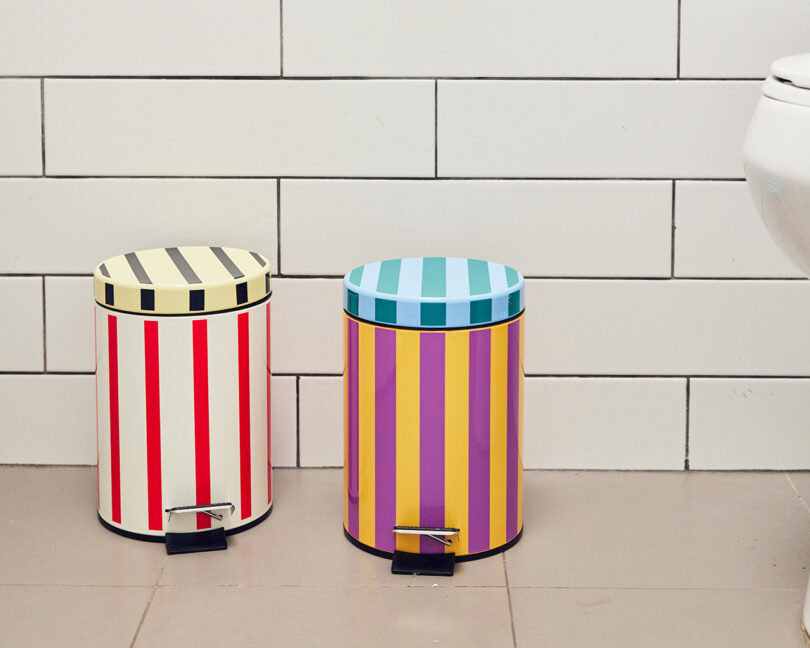Two pedal bins with vertical stripes in different colors are placed on a tiled bathroom floor next to a toilet and a white tiled wall.