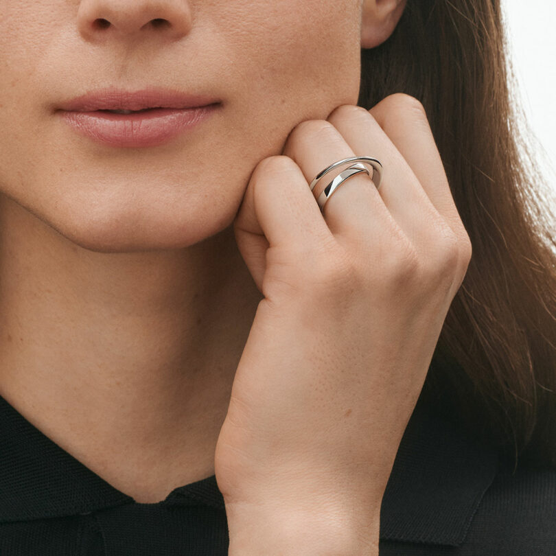 A person with light skin and brown hair rests their chin on their hand, displaying a silver interlocking ring on their finger.