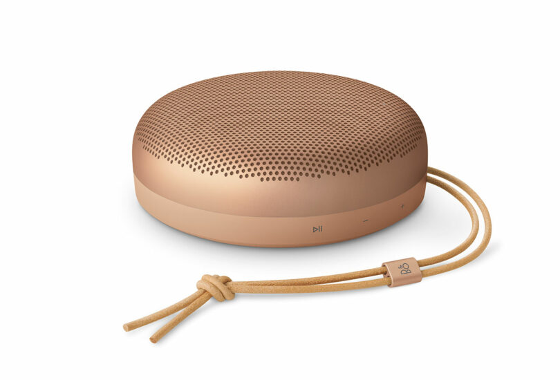 A round, rose gold portable Bluetooth speaker with a perforated top, leather carrying strap, and touch controls—an ideal pick for any Gift Guide.
