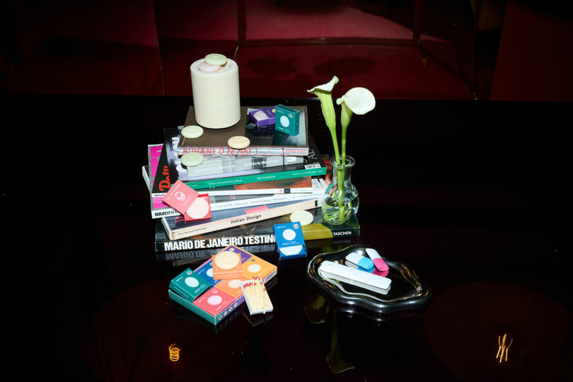 A stack of books, a candle, scattered small colorful boxes, and tealight candles sit on a dark table beside a vase with two white flowers—perfect inspiration for your next Gift Guide.
