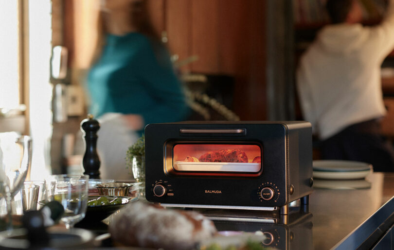 A black BALMUDA toaster oven heating food on a kitchen counter with plates, utensils, and two people in the background.