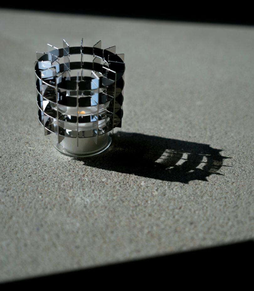 A small cylindrical metal object with a geometric, layered design casts a distinct shadow on a textured surface in natural light.