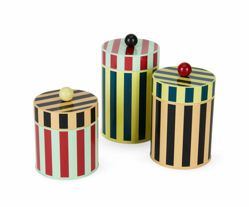 Three cylindrical tins with vertical stripes in different color combinations and round knobs on their lids are displayed against a white background, making them charming gifts for cooks.
