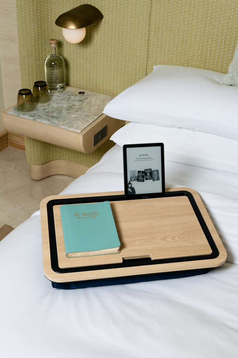 A wooden lap desk on a bed holds a closed teal book and an e-reader displaying a black-and-white page, blending traditional reading with modern technology. A bedside table with glasses, bottles, and a lamp sits in the background.