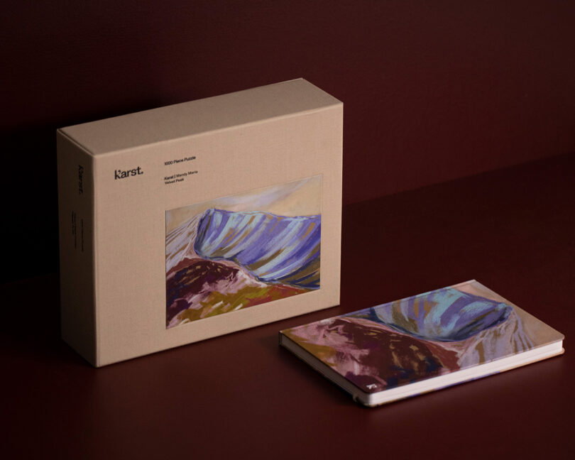 A boxed puzzle and a closed notebook, both featuring abstract mountain artwork, are displayed on a dark brown surface.