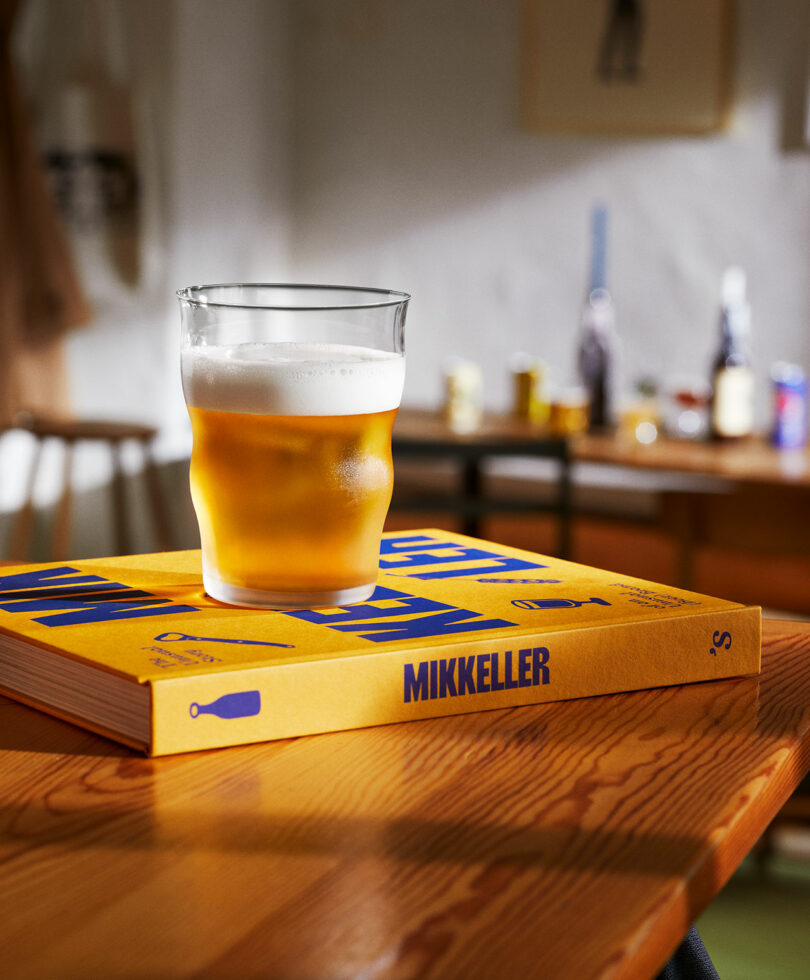 A glass of beer with a foamy head sits on top of a yellow book titled "Mikkeller" on a wooden table, with a blurred background of bottles and objects.