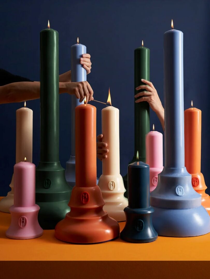 Several hands light tall and short sculpted candles in various colors on an orange surface, with a dark blue background.