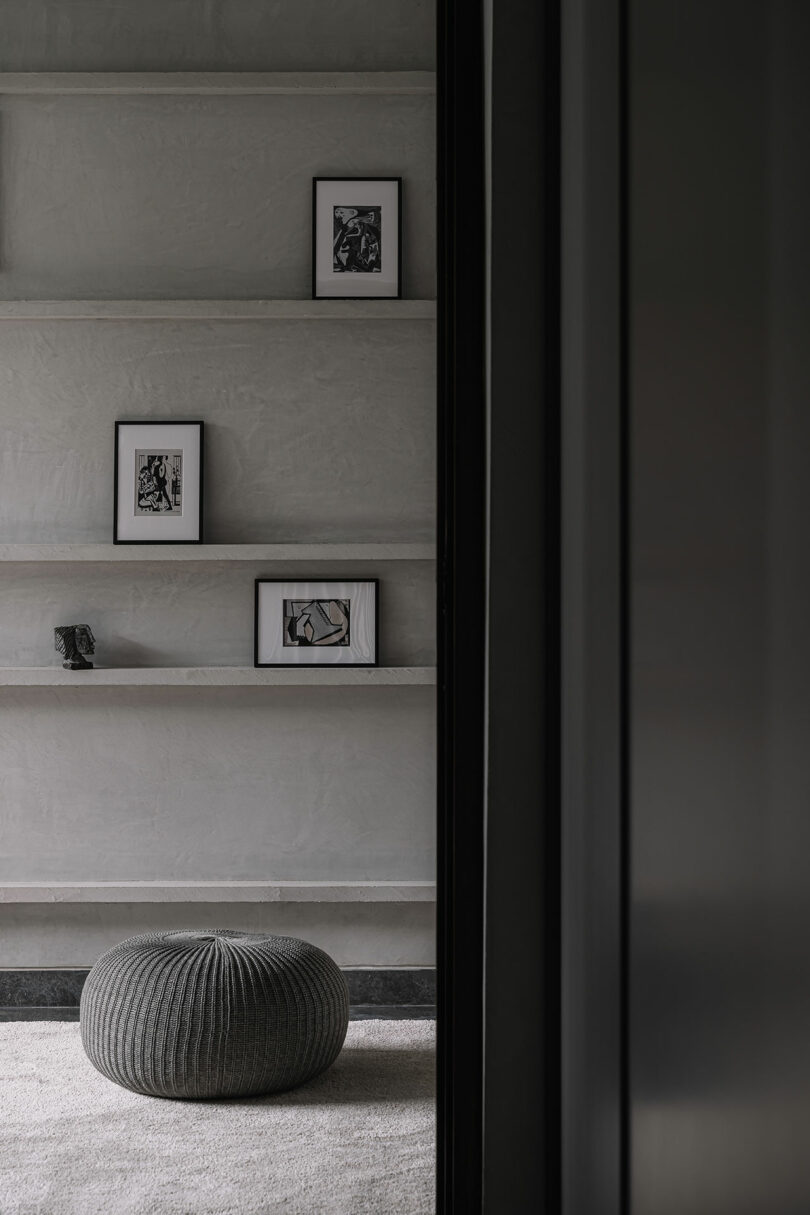 Minimalist room with concrete shelves displaying three framed abstract artworks and a small sculpture, with a round knitted pouf on a carpeted floor in the foreground.