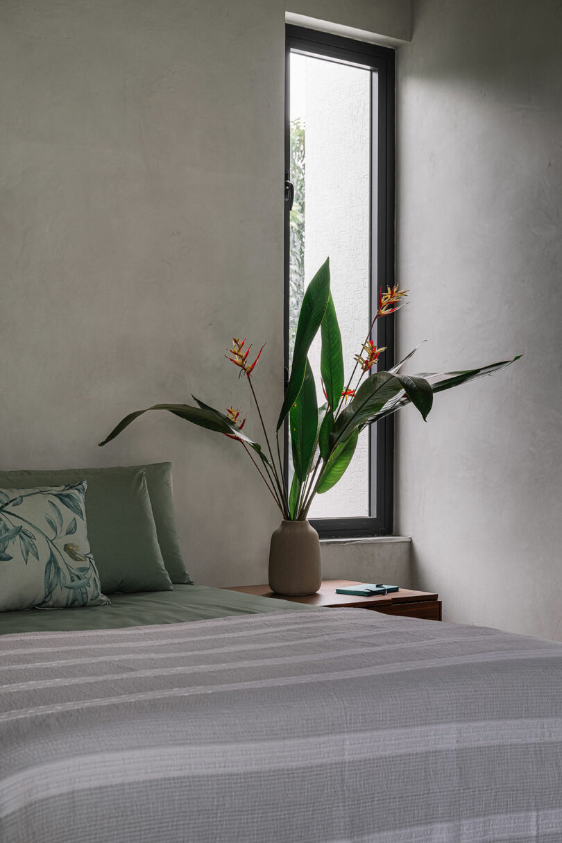 A neatly made bed with a striped white and gray cover, green pillows, a bedside table with a vase of tropical flowers, and a window letting in natural light.