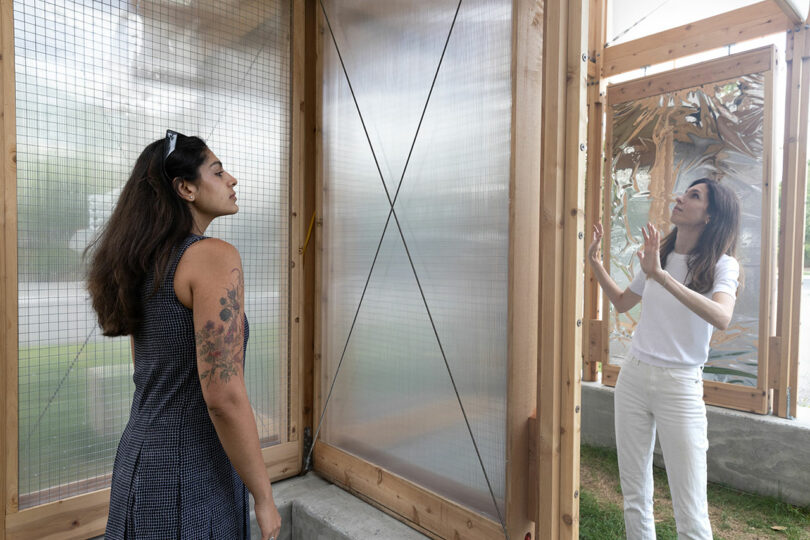 Two women stand inside a wooden and translucent panel structure, one observing while the other gestures and explains.