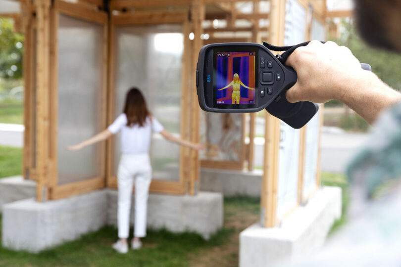 A person uses a thermal camera to capture the heat signature of a woman standing with arms outstretched inside a wooden and plastic structure outdoors.