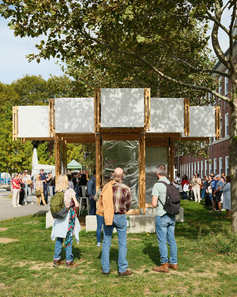 A group of people gather outdoors around a modern wooden pavilion with large glass panels and white rectangular features on a grassy area near trees and buildings.