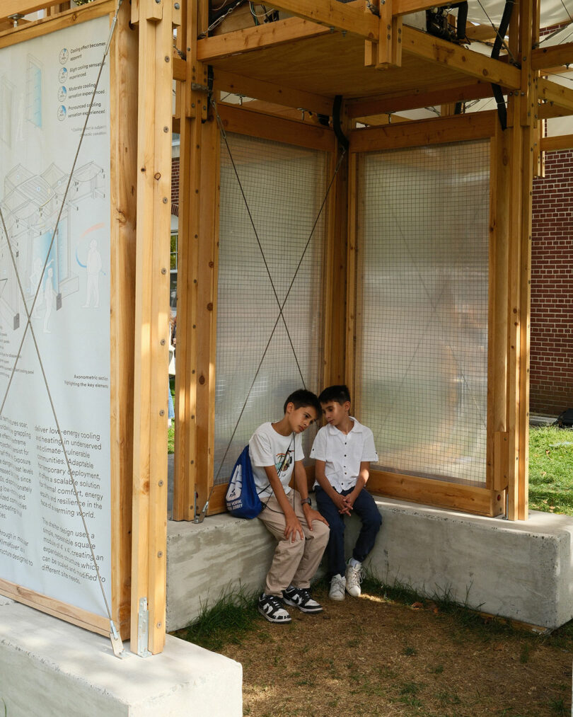 Two children sit closely together on a concrete bench under a wooden structure, with a diagram and brick building visible in the background.