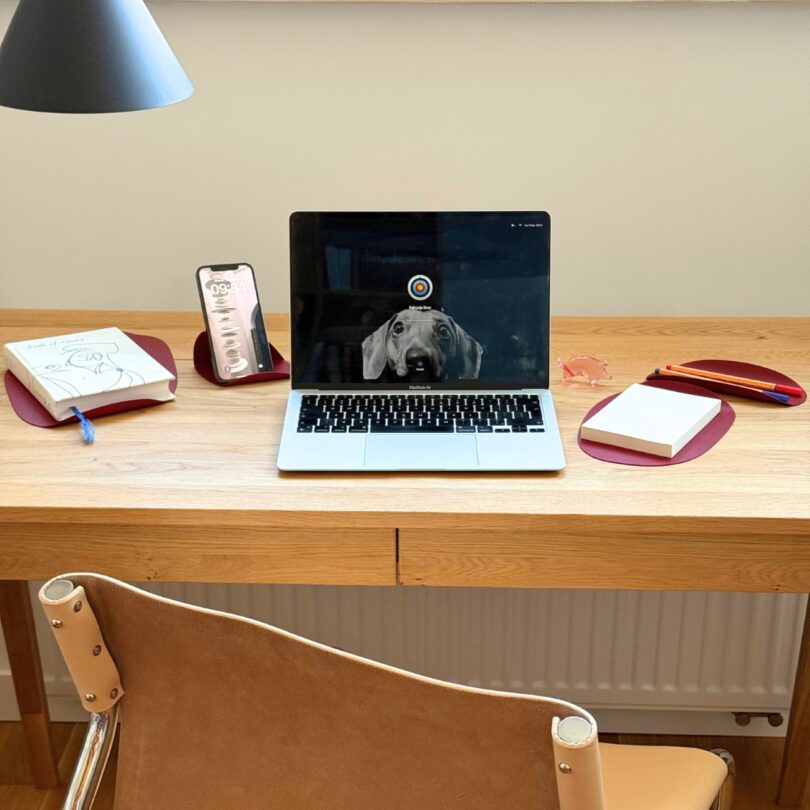 A wooden desk with a laptop displaying a dog, a phone, notebook, pens, sticky notes, and a lamp; a tan chair is positioned in front of the desk