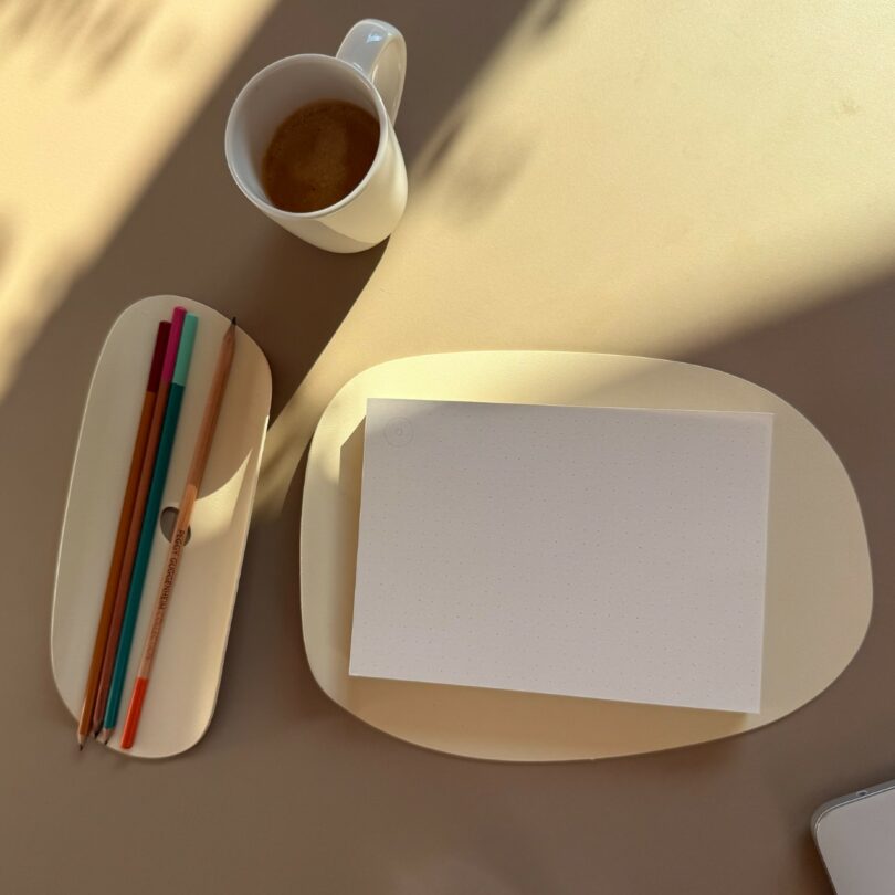A cup of coffee, three colored pencils, a pen, and a blank dotted sheet of paper on cream-colored trays sit on a beige surface in natural light