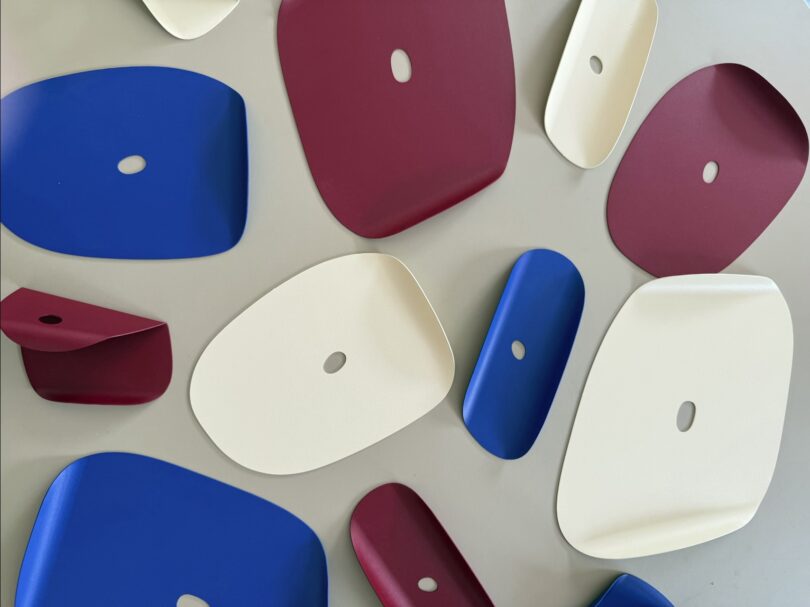 Several flat, rounded geometric trays in maroon, blue, and cream colors, each with a central oval hole, are arranged on a light gray surface.