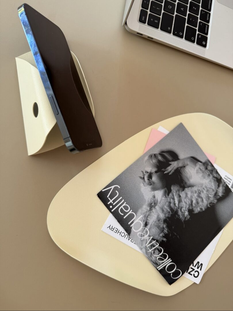 A smartphone on a stand, a laptop, and a tray with magazines and a black-and-white photograph on a beige surface