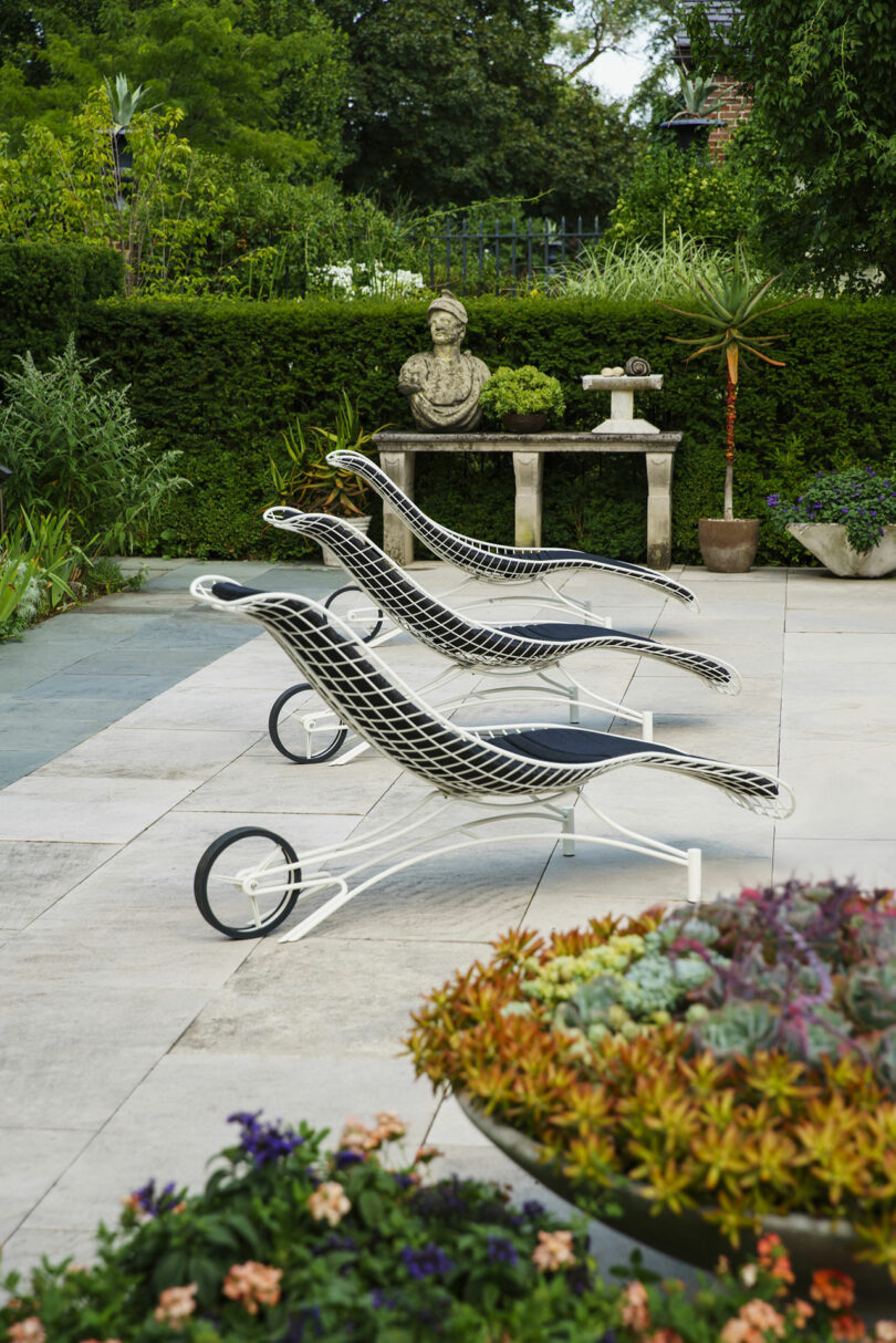 Three modern black and white lounge chairs are arranged on a patio near a table with a bust sculpture, surrounded by lush greenery and potted plants.