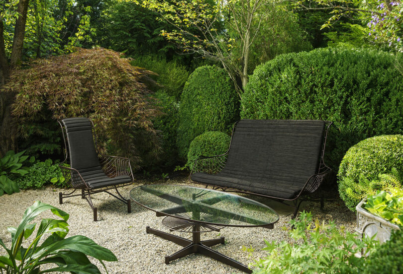 Metal patio furniture, including a chair, a bench, and a glass-top table, is set on a gravel area surrounded by lush green shrubs and trees in a garden.