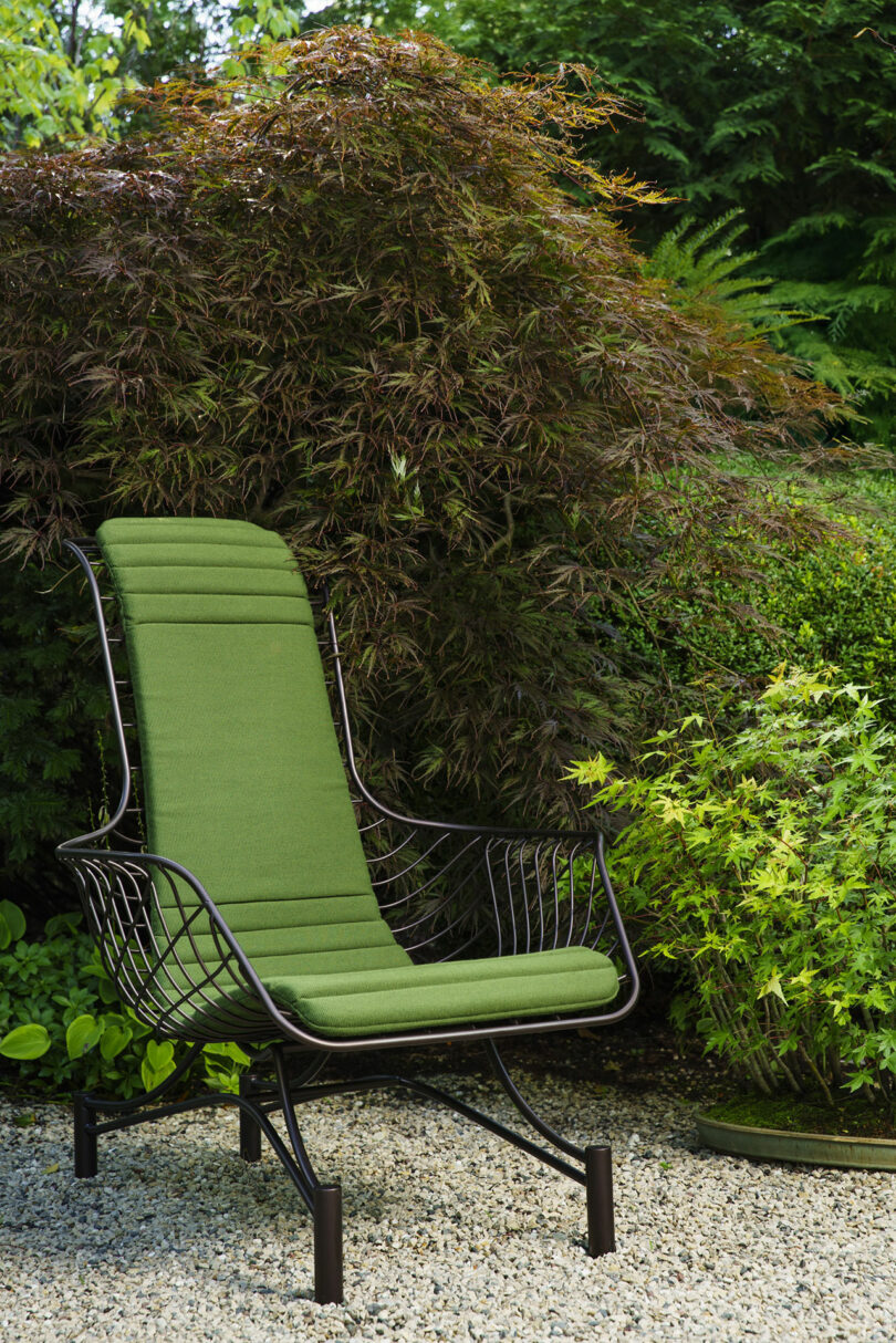 A green cushioned metal chair sits on a gravel surface in a garden, surrounded by various leafy bushes and plants.