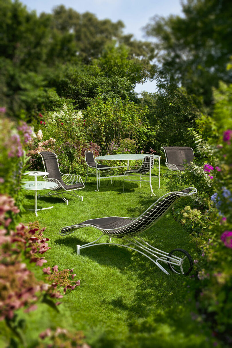 A set of black and white outdoor lounge chairs and a table are arranged on a grassy lawn surrounded by lush green plants and colorful flowers.