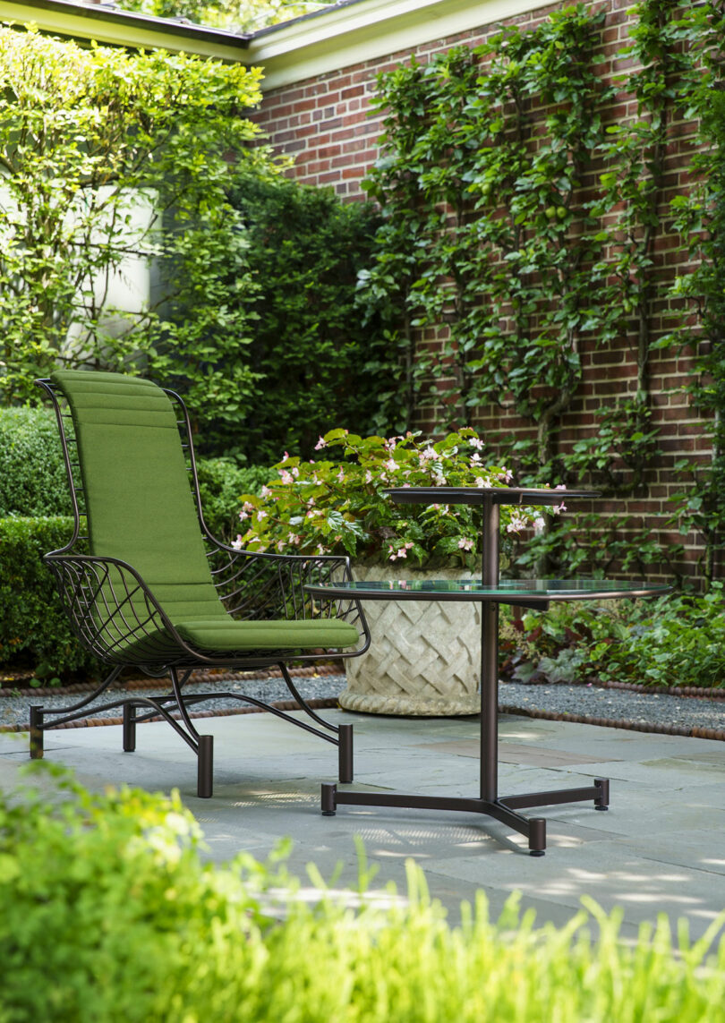 A green cushioned chair and a small round black table sit on a stone patio, surrounded by lush greenery and a brick wall covered in climbing plants.