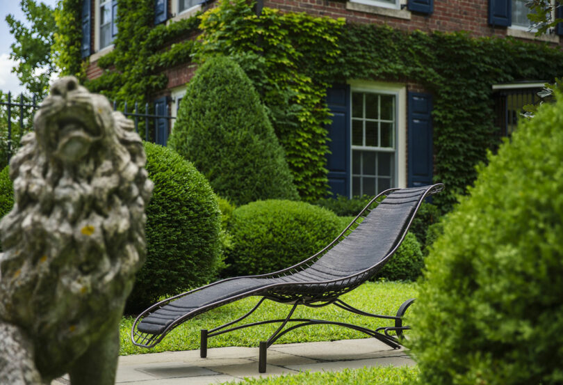 A black metal lounge chair sits on a stone patio surrounded by manicured bushes, topiaries, a stone lion statue, and a brick house covered in ivy.