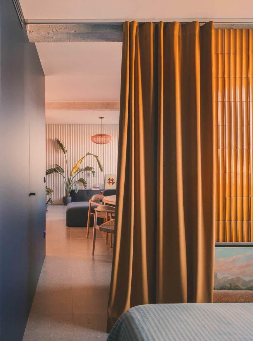 A modern interior with a gold curtain partially dividing a bedroom from a living and dining area, featuring plants, wood accents, and soft lighting.