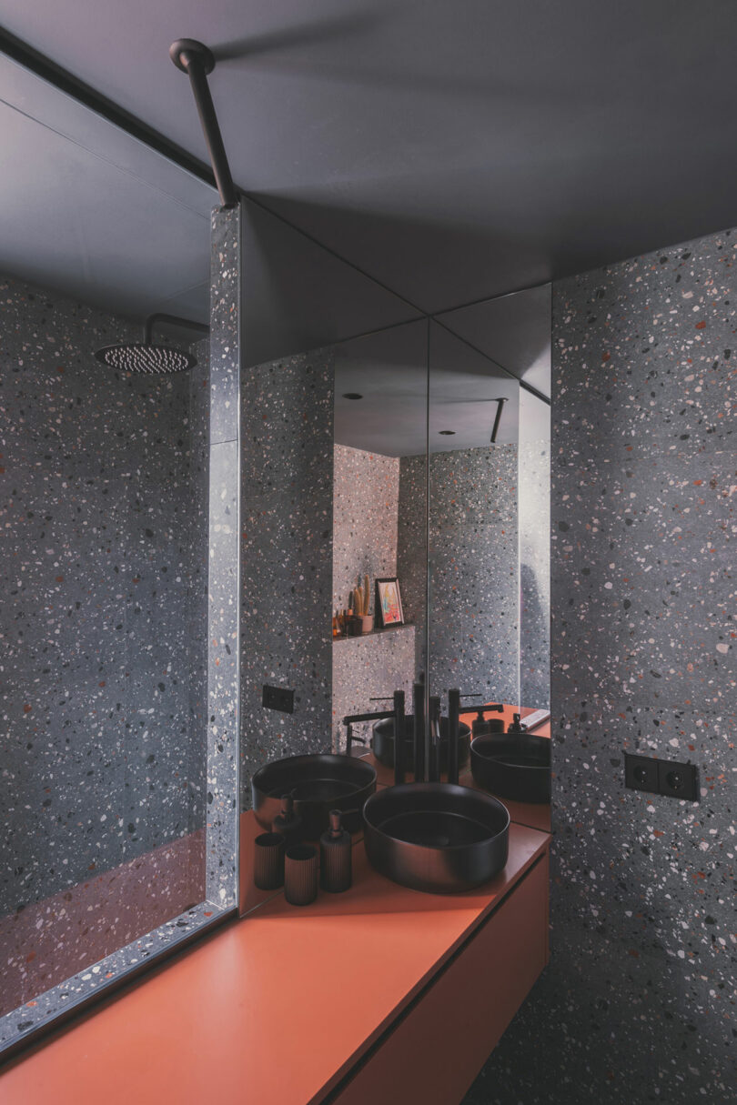Modern bathroom with terrazzo walls, a large mirror, black sink and fixtures, and an orange countertop. A rainfall showerhead is reflected in the mirror.