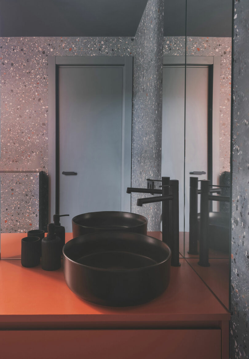 A modern bathroom with a terrazzo wall, a round black vessel sink, matte black faucet, black accessories, and a mirrored cabinet above a coral countertop.