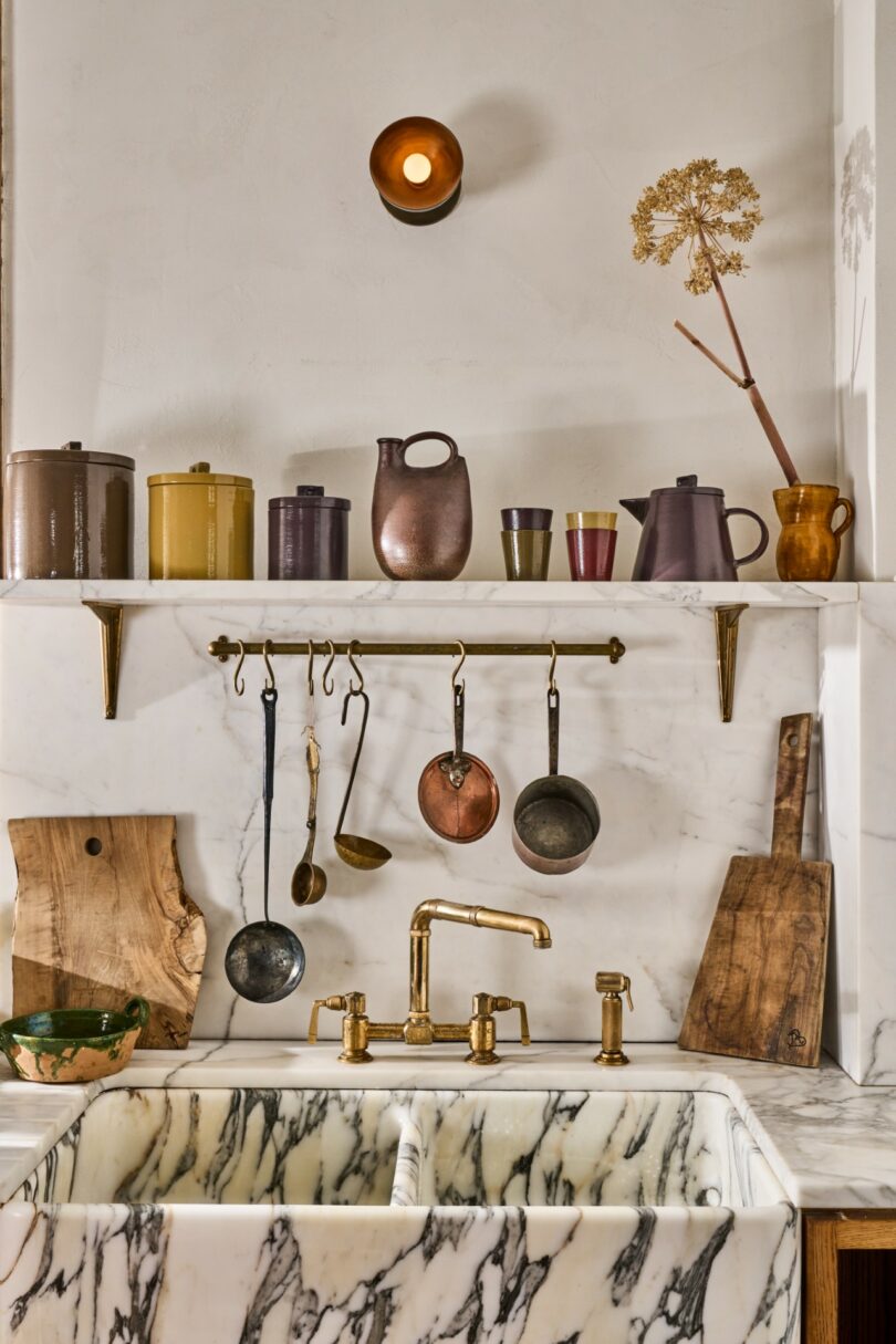 A marble kitchen sink with a brass faucet, wooden cutting boards, utensils hanging above, and ceramic jars and cups arranged on a shelf against a white wall