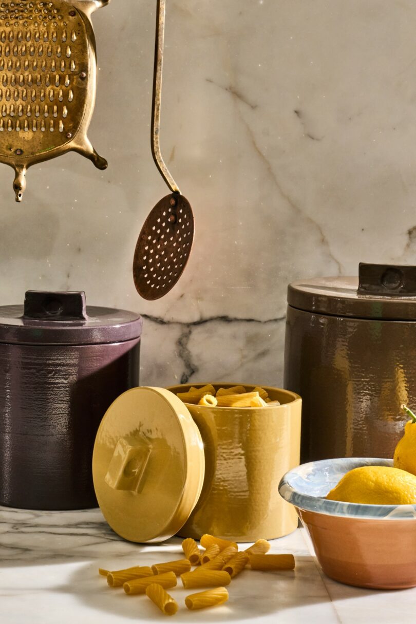 Three metal canisters, one open with pasta inside, sit on a marble countertop beside a metal bowl with lemons, with brass kitchen utensils hanging above