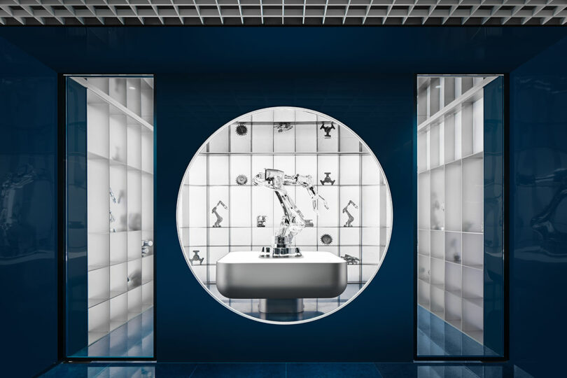 A robotic arm displayed in a circular window set in a blue wall, surrounded by white-lit shelves and a tiled background featuring small robot illustrations.