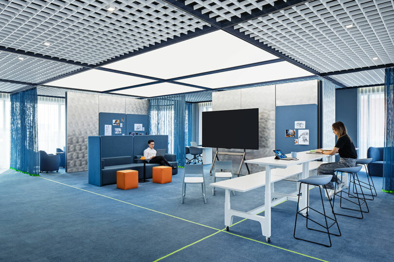 Modern office space with blue carpet, modular seating, tables, stools, and two people working on laptops. Large screen and whiteboard are visible in the center of the room.