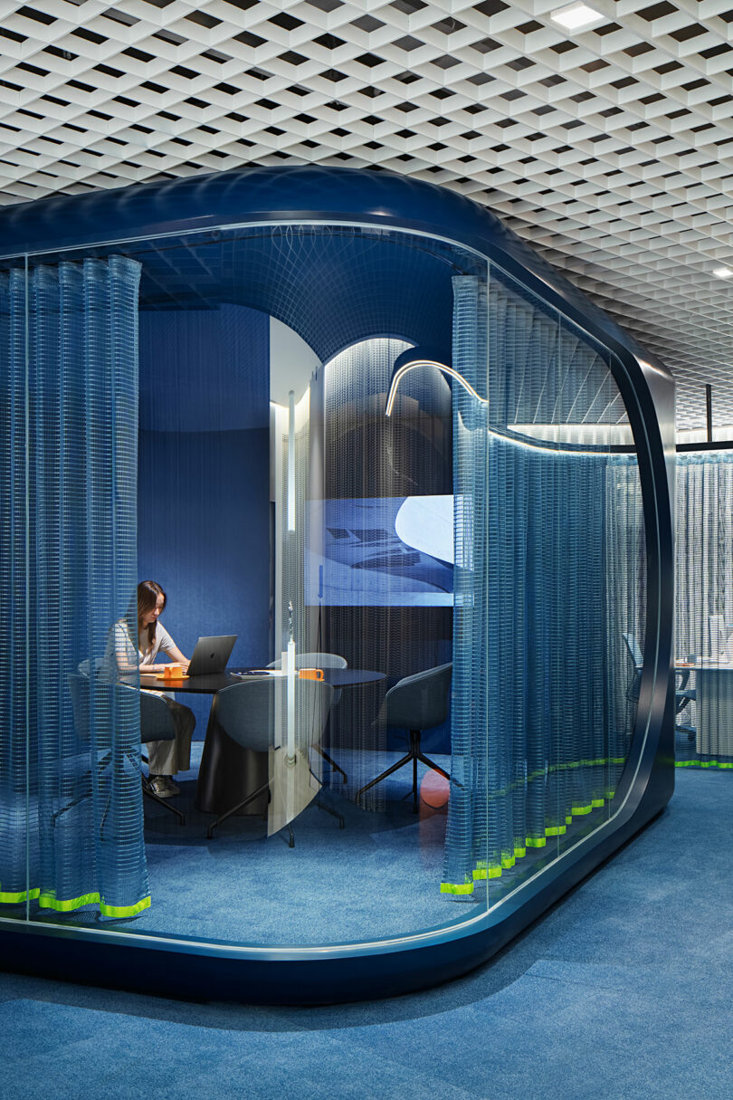 A person works at a desk with a laptop in a modern, glass-walled office pod with blue carpeting and curved blue curtains.