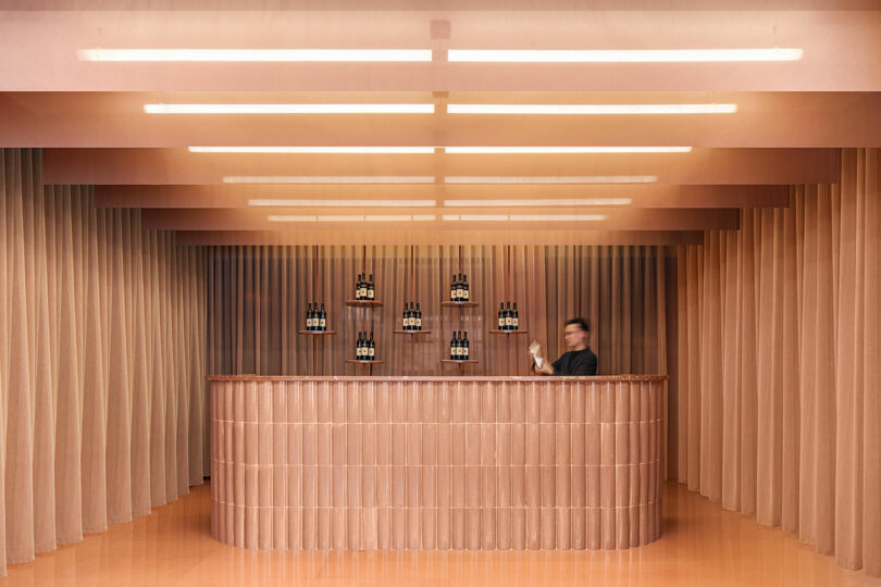 A bartender stands behind a curved, textured bar in a minimalist room with warm lighting, beige curtains, and shelves displaying bottles on the back wall.