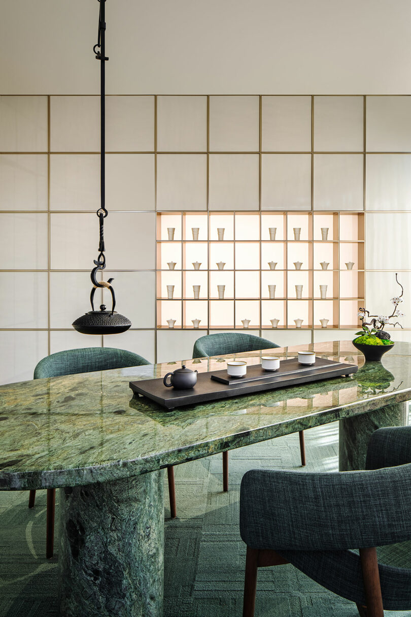 A modern dining area with a green marble table, dark upholstered chairs, a black hanging light, and a minimalist display of ceramic cups on a backlit wall shelf.