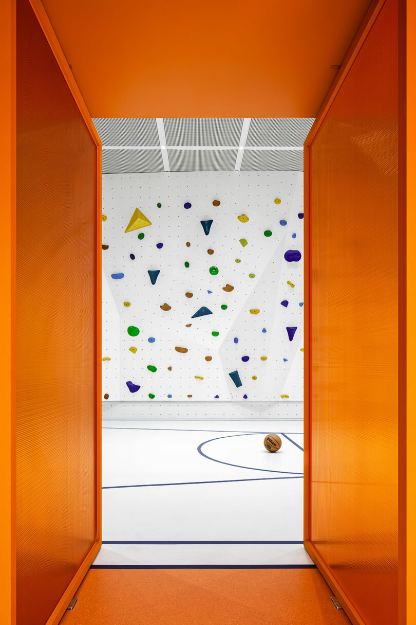 View through orange double doors into a gym with a white climbing wall featuring colorful holds and a basketball lying on a white court with blue lines.