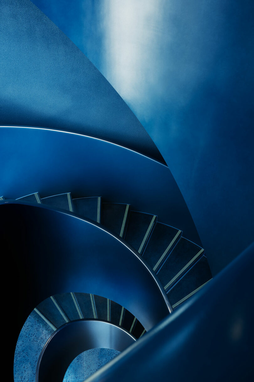 A spiral staircase with blue steps and walls, viewed from above, creating a smooth circular pattern and highlighting the modern architectural design.