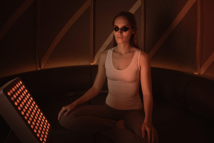 A person wearing protective goggles sits cross-legged on a couch, illuminated by a red light therapy panel in a dimly lit room.
