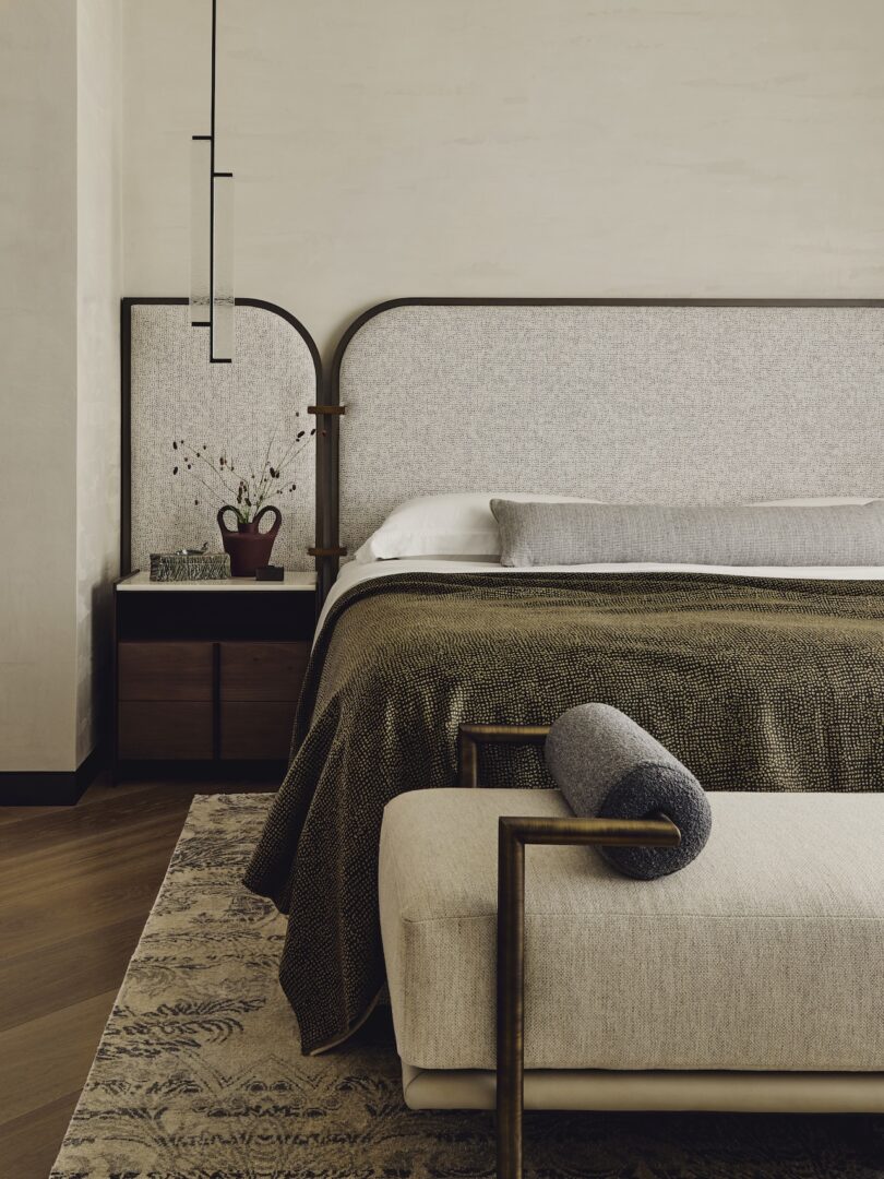 A modern bedroom features a large bed with a textured headboard, brown blanket, and gray accent pillow, next to a dark wood nightstand and a cushioned bench at the foot of the bed.