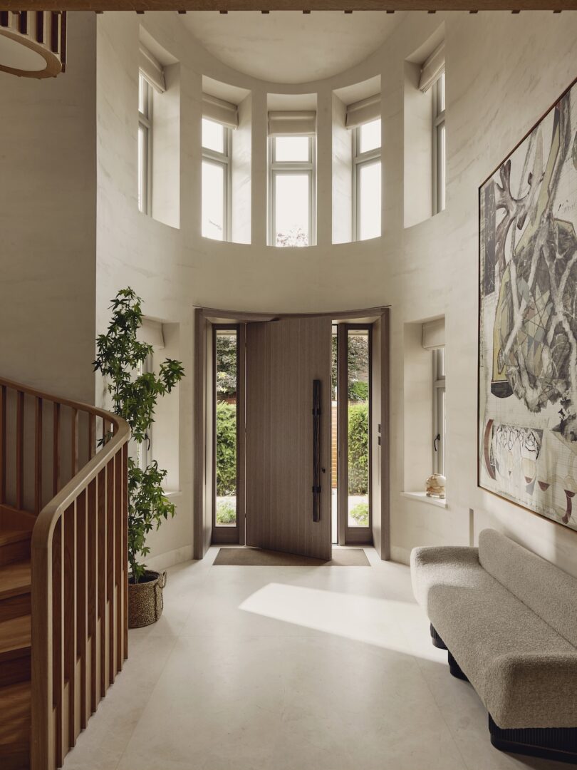 Modern entryway with tall windows, wooden staircase, potted plant, large abstract painting, curved sofa, and double doors opening to a garden.