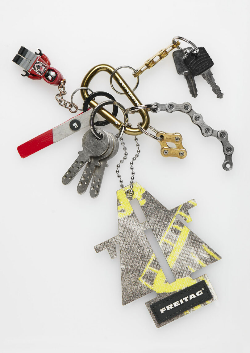 A set of various keychains and keys, including a toy figure, a red and white stick, metal keys, a chain link, and a Freitag-branded tag, all attached to a gold carabiner.