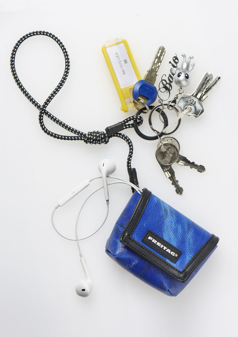 A set of keys with various keychains, a yellow USB stick, and a blue FREITAG pouch with white earphones are arranged on a white surface.
