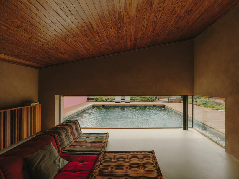 Interior view of a living room with a wooden ceiling and large window overlooking an outdoor swimming pool and two white lounge chairs.