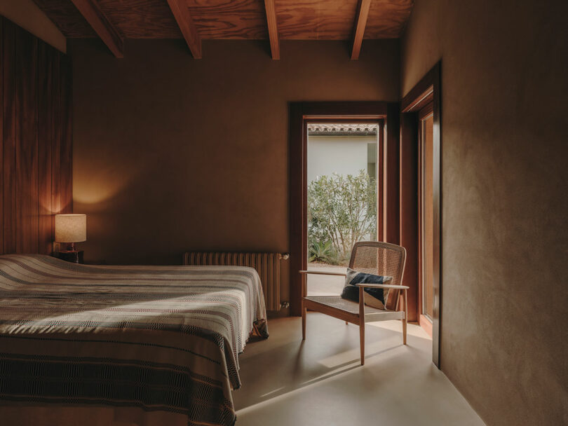 A minimalist bedroom with a bed, striped bedding, a wooden chair, a lamp on a nightstand, and an open door leading to a courtyard with greenery.
