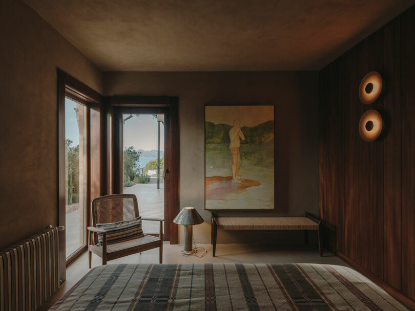 A warmly lit bedroom with a bed, a chair, a woven bench, a lamp, a large painting on the wall, and a glass door leading to an outdoor patio with a scenic view.