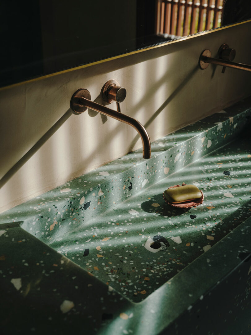 A terrazzo sink with a bar of soap and two wall-mounted bronze faucets is illuminated by sunlight streaming through window blinds.