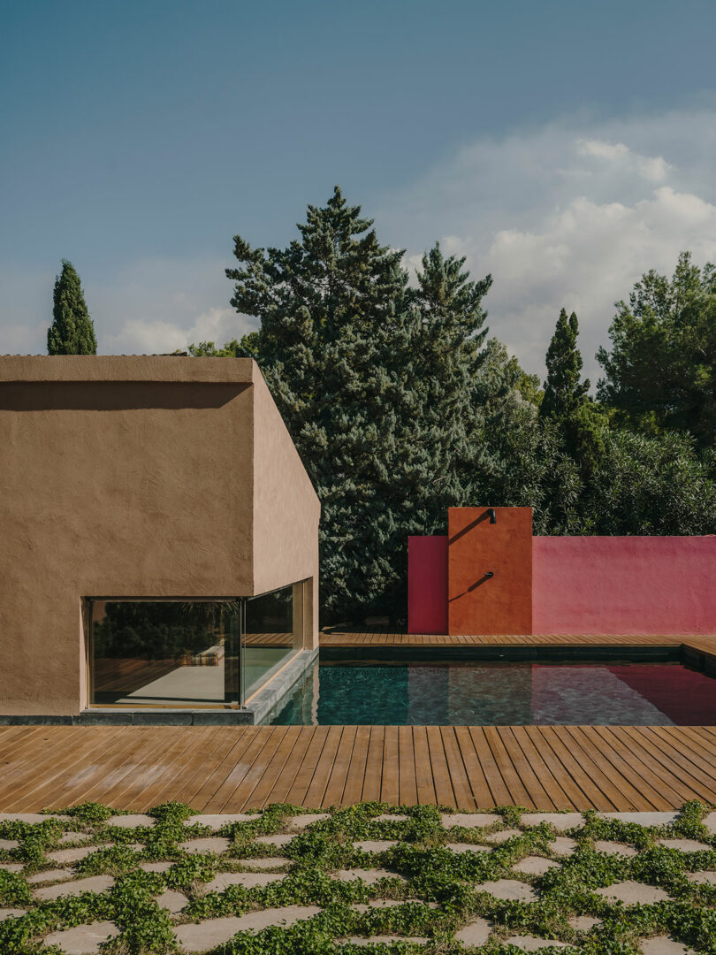 A modern house with textured walls, large windows, a wooden deck, and a small pool, surrounded by greenery and trees, with a pink accent wall in the background.